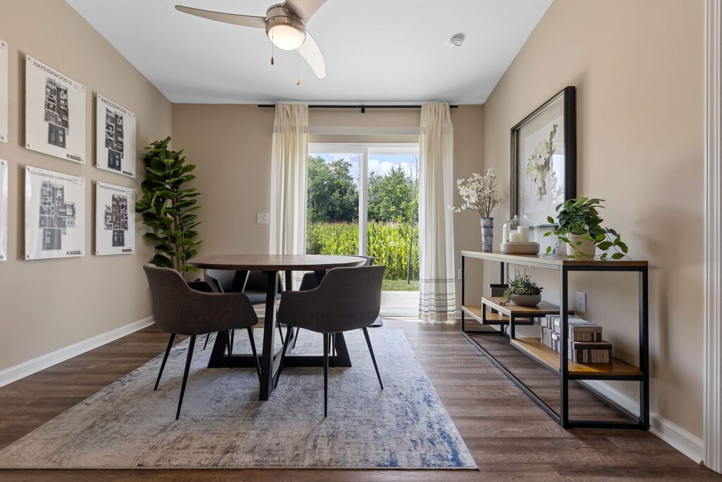 Modern dining room with a round table, four chairs, large window with sheer curtains, wall art, plants, and a console table on wood flooring. Sunlight streams in from outside.