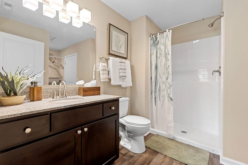 Modern bathroom with a dark wood vanity, granite countertop, toilet, shower with a white curtain, framed artwork, and neutral-colored walls and flooring.