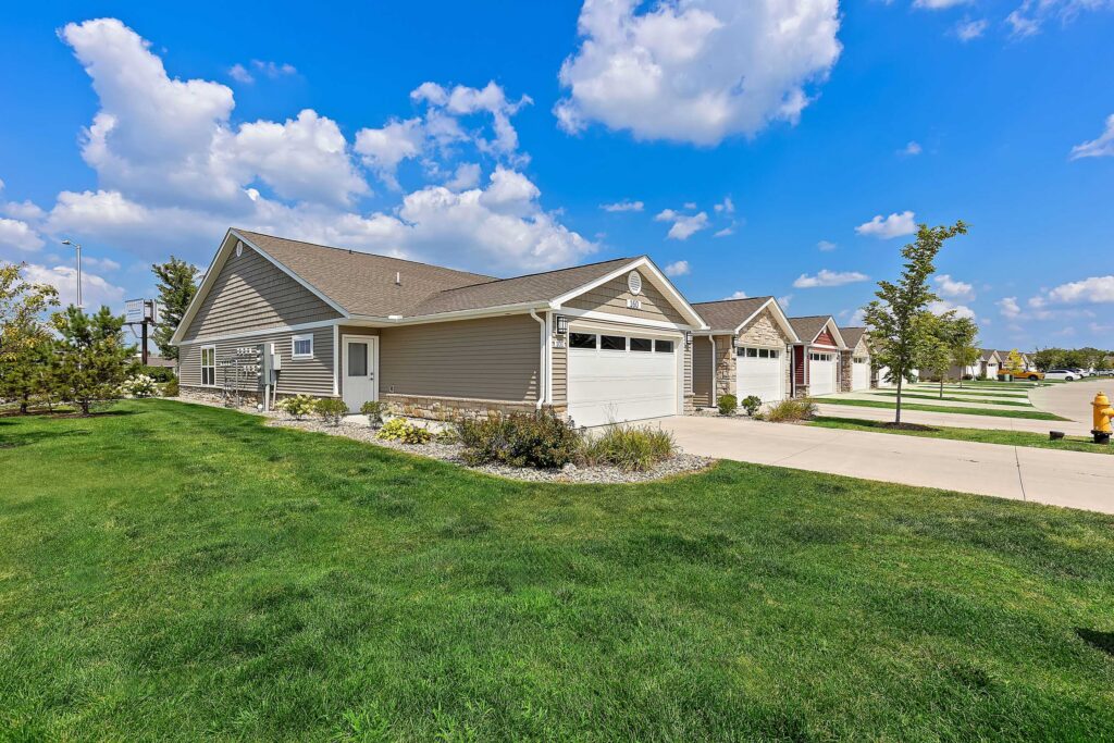 Single-story suburban house with attached two-car garage, beige siding, and a well-maintained lawn under a blue sky with scattered clouds.