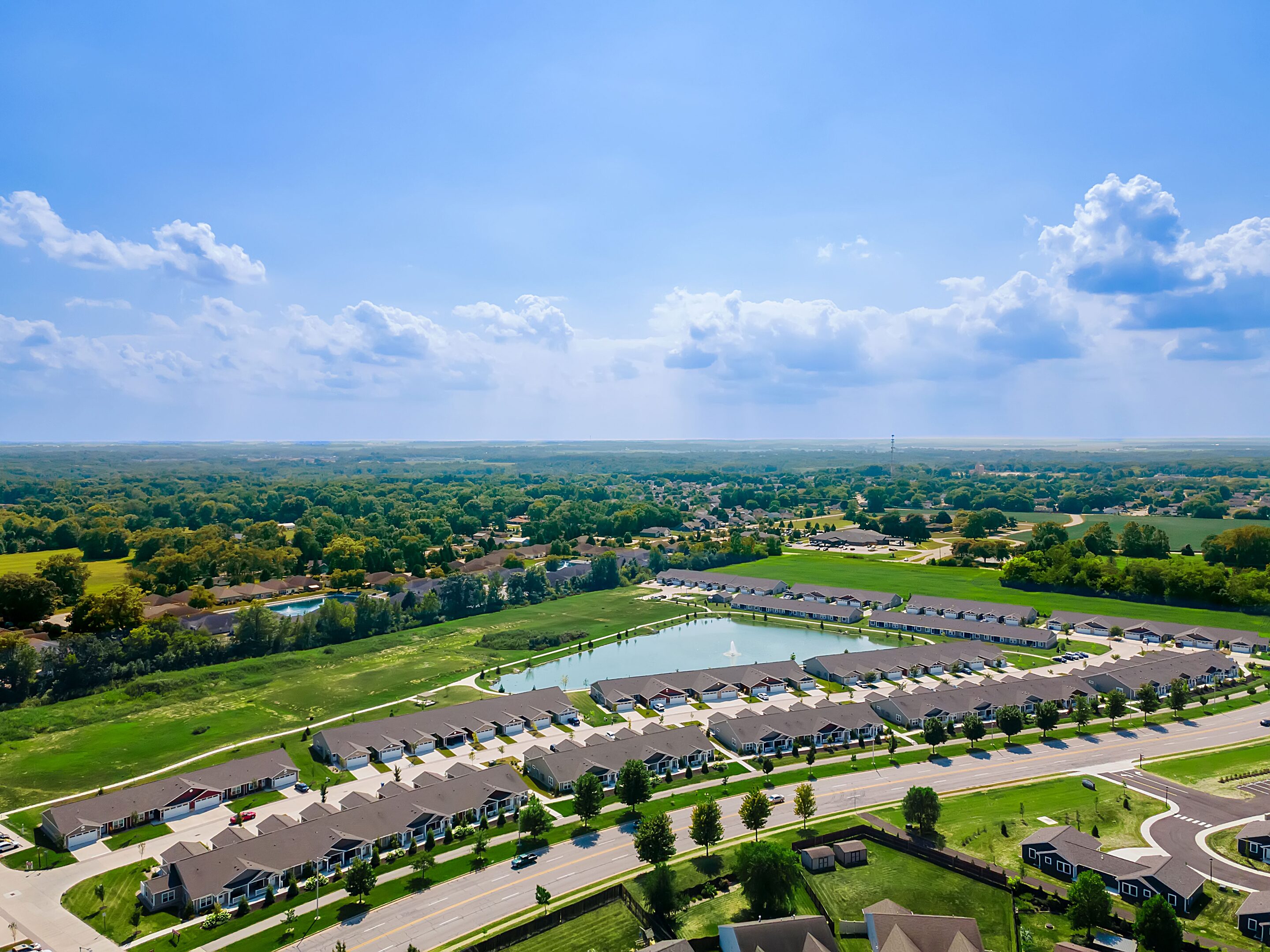 Aerial view of a suburban neighborhood with rows of houses, a pond, green lawns, and surrounding trees under a blue sky with scattered clouds.