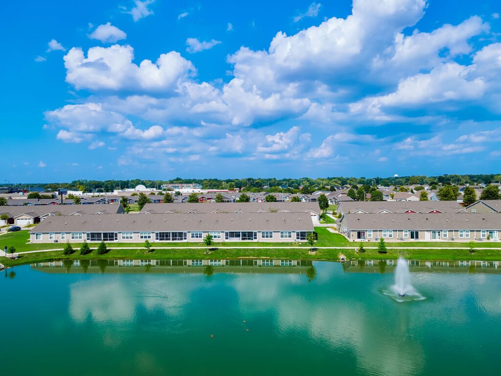 A row of residential buildings lines a pond with a fountain, under a blue sky with scattered clouds. Trees and green lawns surround the water.