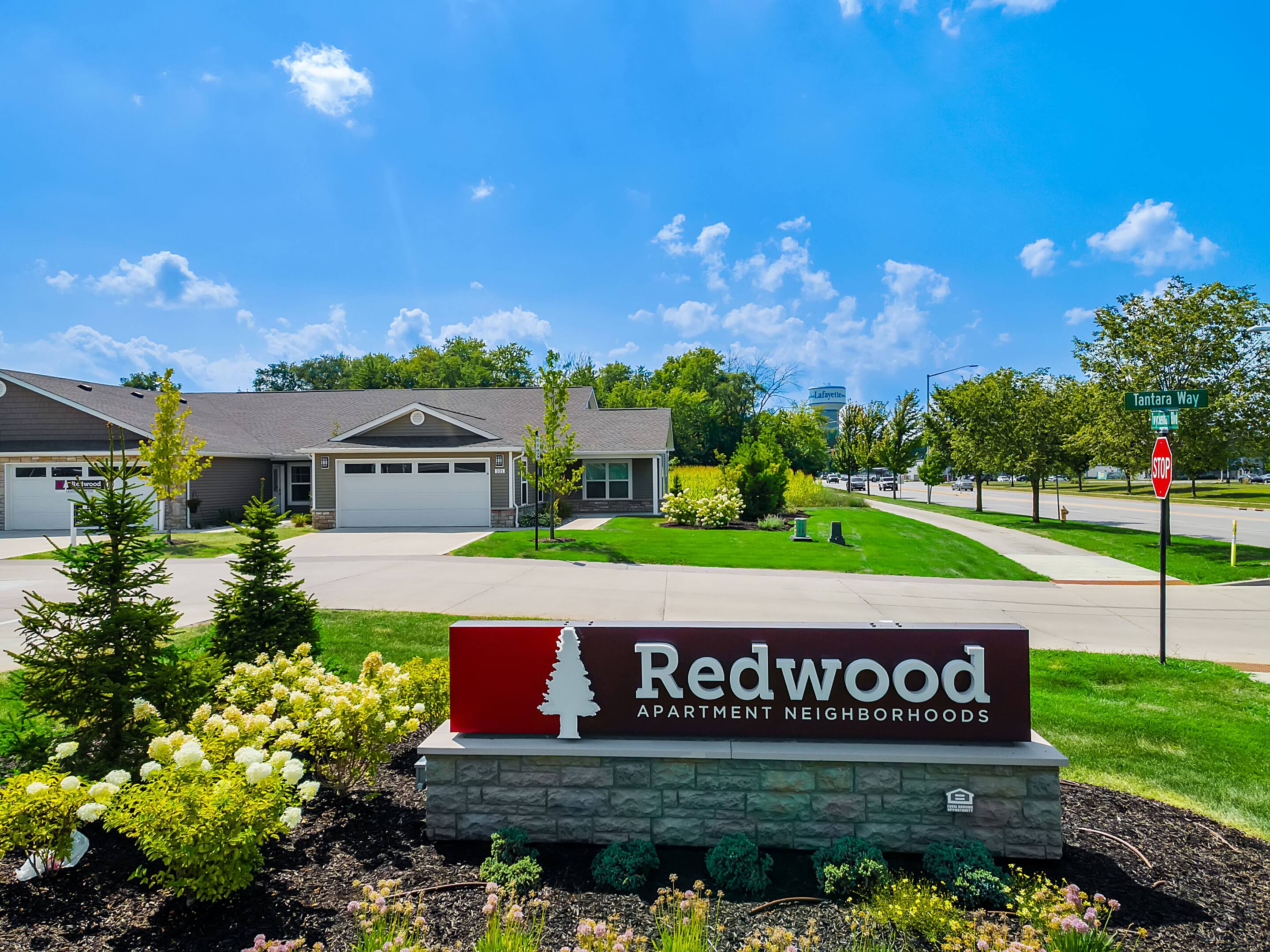 A red and white sign reading "Redwood Apartment Neighborhoods" stands in front of single-story apartments with garages, green lawns, and a clear blue sky.