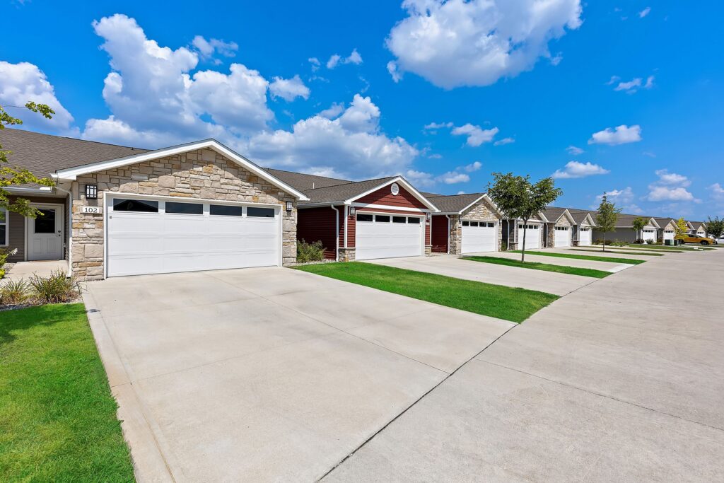 Row of single-story suburban houses with attached garages, neatly mowed lawns, and a wide, clean street under a partly cloudy blue sky.