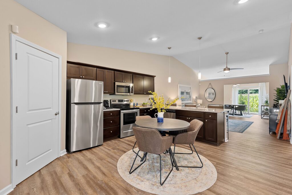 Open kitchen and dining area with stainless steel appliances, dark wood cabinets, a round table with four chairs, and a view into the living room and patio.
