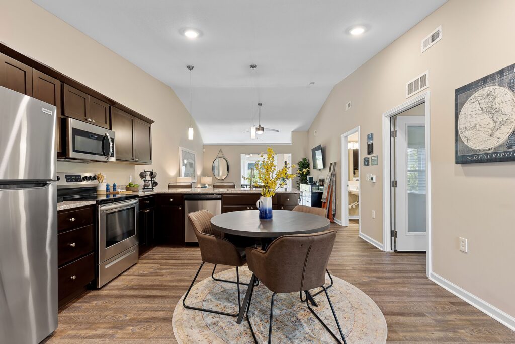 Modern kitchen and dining area with dark wood cabinets, stainless steel appliances, round table with four chairs, and decorative items on light wood flooring.