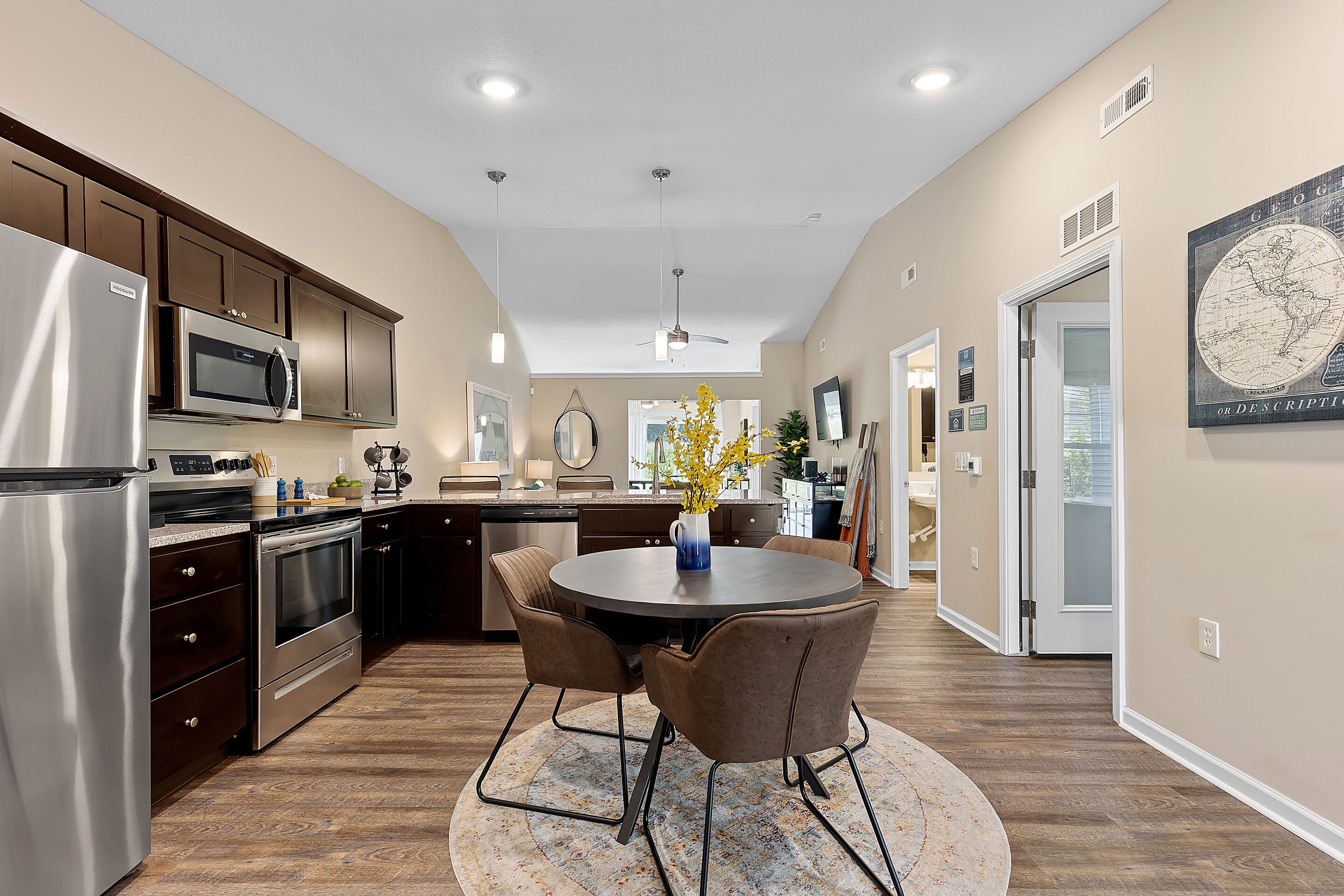 Modern kitchen and dining area with dark wood cabinets, stainless steel appliances, round table with four chairs, and decorative items on light wood flooring.