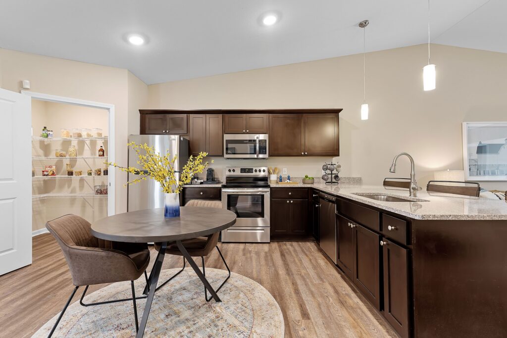 Modern kitchen with dark cabinets, stainless steel appliances, a round dining table with two chairs, and a walk-in pantry visible through an open door.
