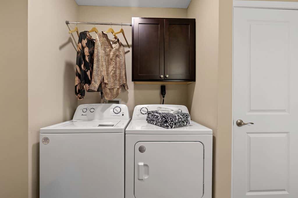 A laundry room with a washing machine and dryer side by side, shirts hanging on a rod above, a cabinet mounted on the wall, and a laundry basket on the dryer.