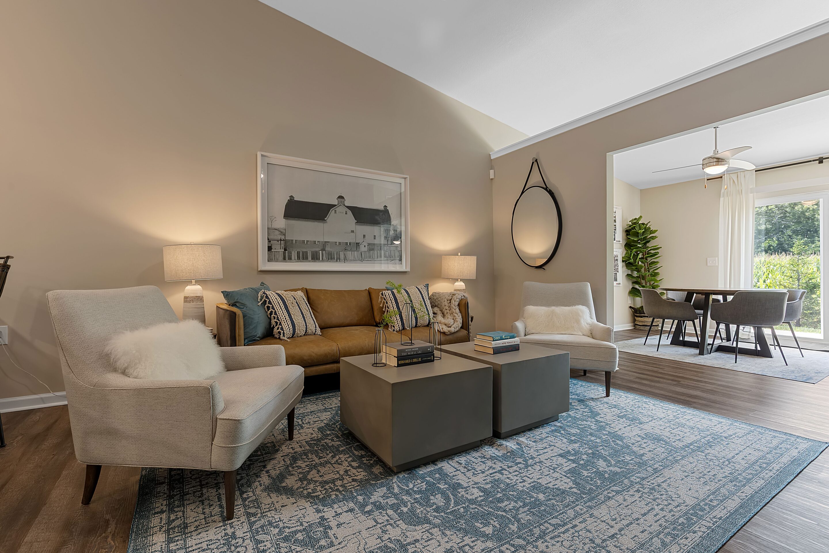 Modern living room with a tan sofa, two beige armchairs, a blue patterned rug, two cube coffee tables, wall art, and a dining area visible through an open doorway.