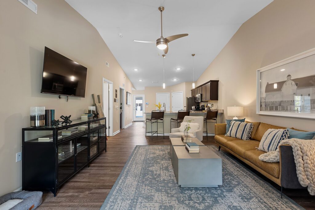 A modern living room with a wall-mounted TV, glass console, coffee table, beige sofa, blue rug, and open view of a kitchen and dining area in the background.