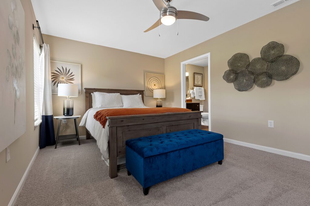 A bedroom with a wooden bed, white bedding, orange blanket, blue upholstered bench, beige walls, two lamps, and an open door to a bathroom.