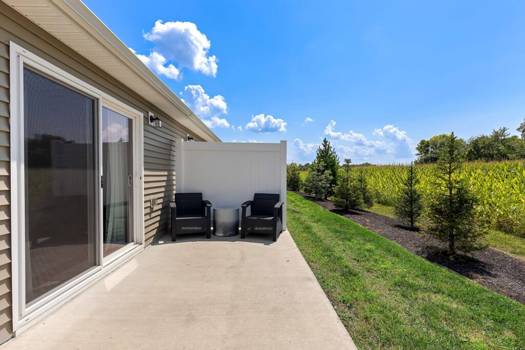 A concrete patio with two black chairs and a small table sits beside a tan building, overlooking a grassy area and cornfield under a blue sky with scattered clouds.
