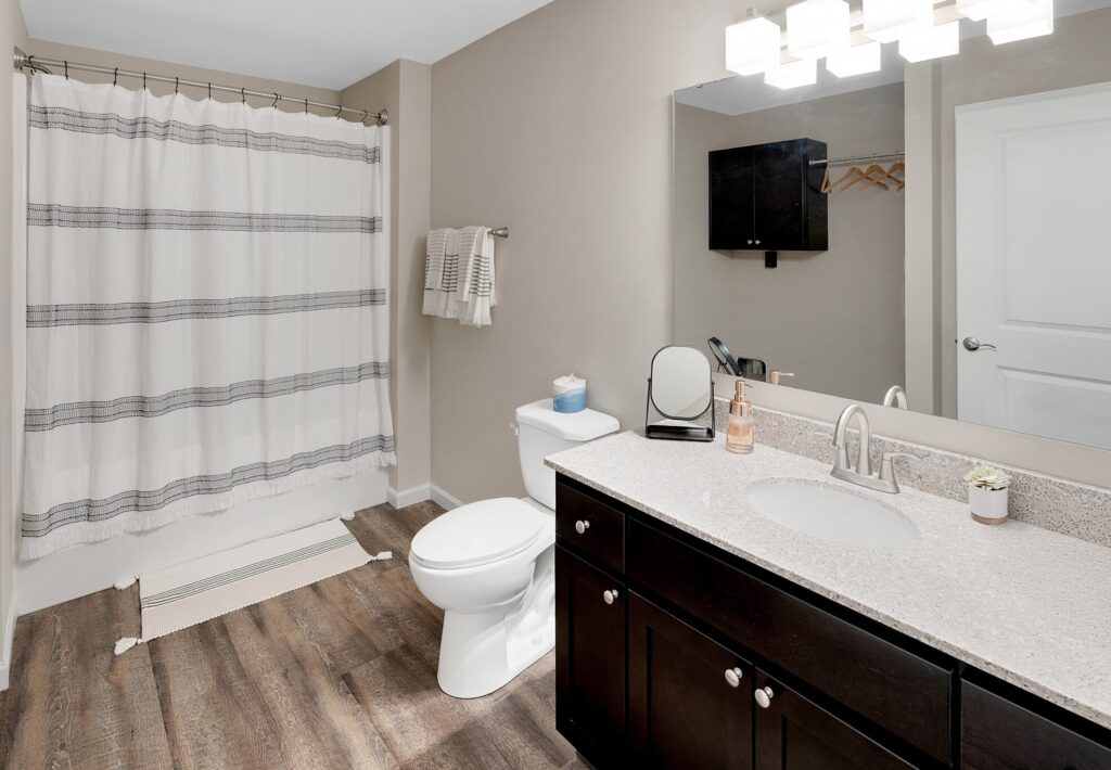 Modern bathroom with a dark wood vanity, white countertop, toilet, striped shower curtain, wall-mounted towels, and a large mirror under bright overhead lights.