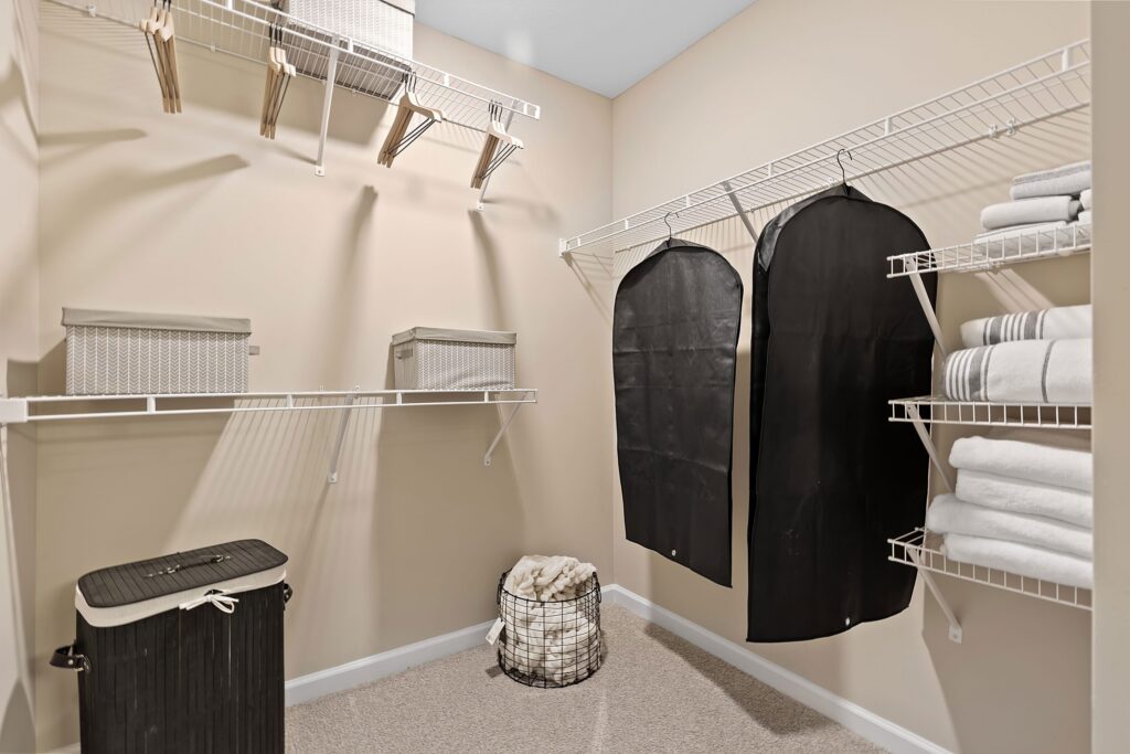 A walk-in closet with wire shelves holding baskets, folded towels, black garment bags, a hamper, and a basket of rolled towels on a carpeted floor.