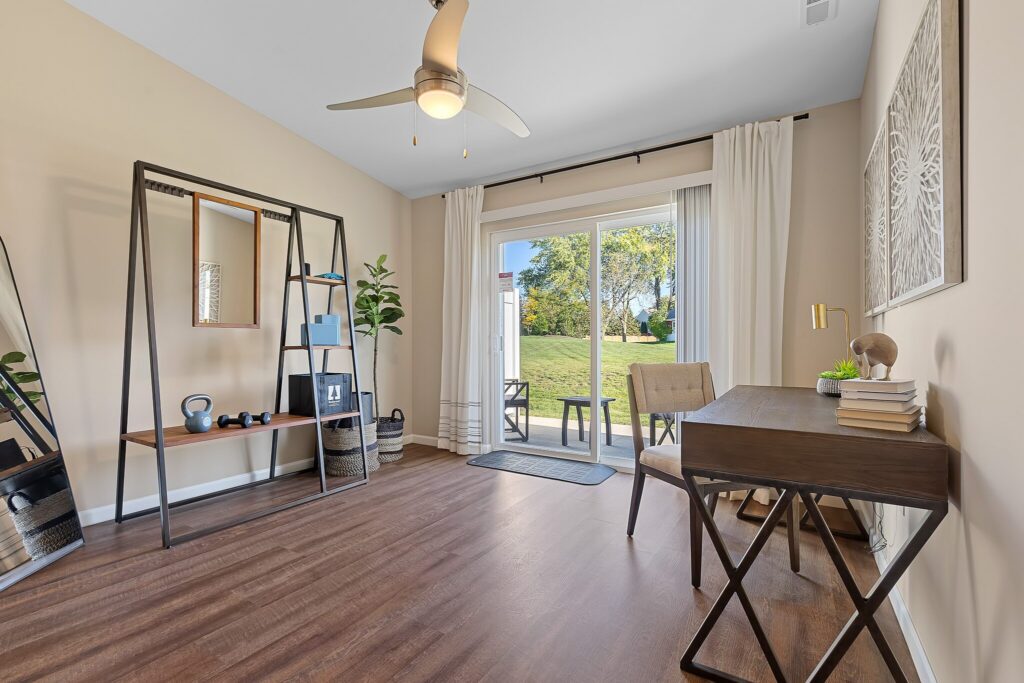 A modern home office with a desk, chair, shelving unit, exercise equipment, and large sliding doors overlooking a grassy backyard.
