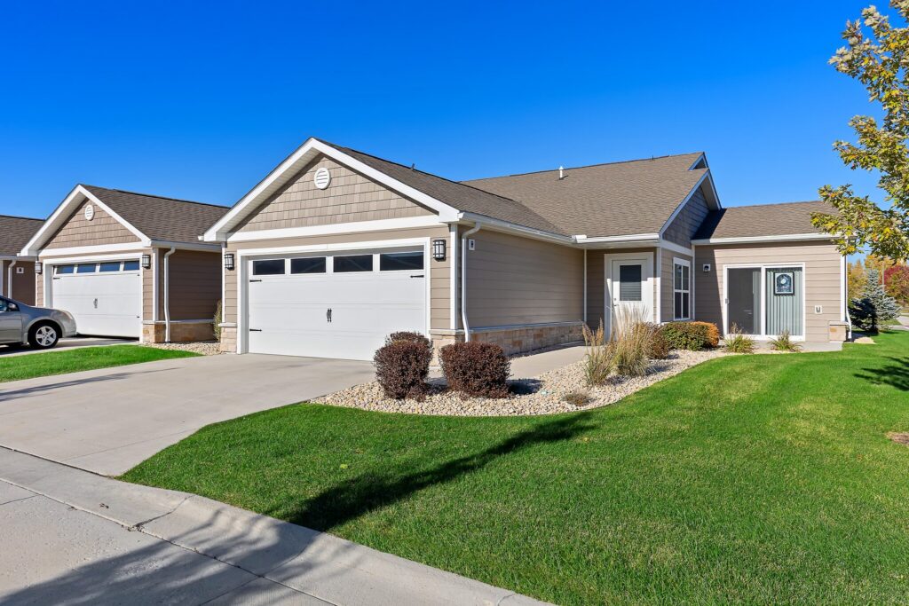 Single-story suburban house with a double garage, beige siding, white trim, a well-maintained lawn, and a concrete driveway on a clear, sunny day.