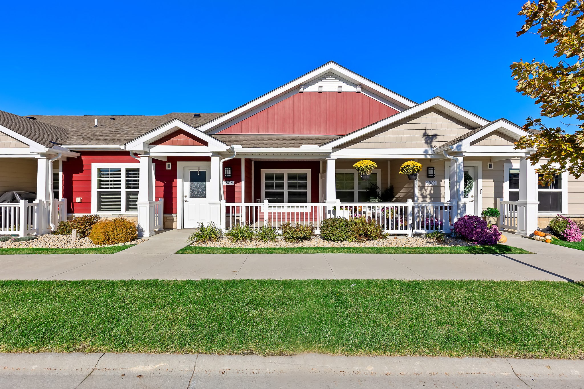 Single-story house with a red and beige exterior, white trim, front porch, manicured lawn, and flower beds under a clear blue sky.