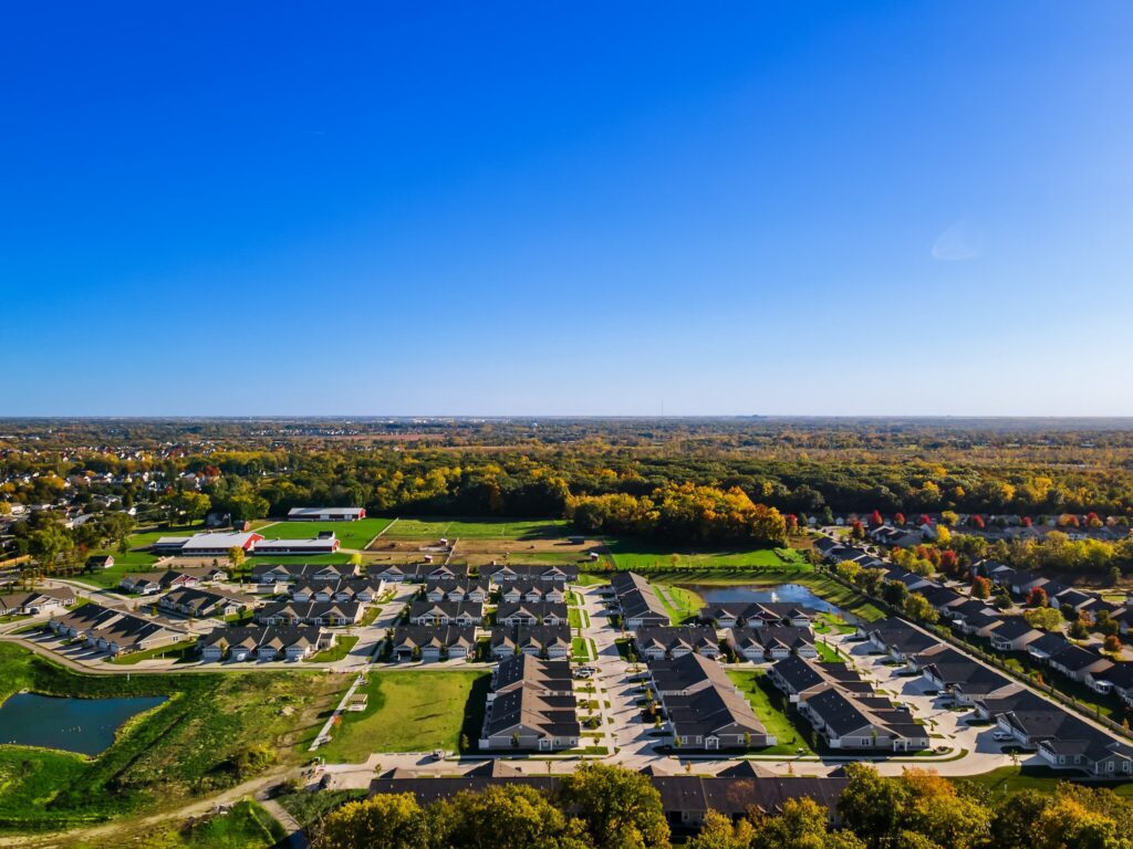 Aerial view of a suburban neighborhood with rows of houses, green spaces, ponds, and a forested area in the background under a clear blue sky.