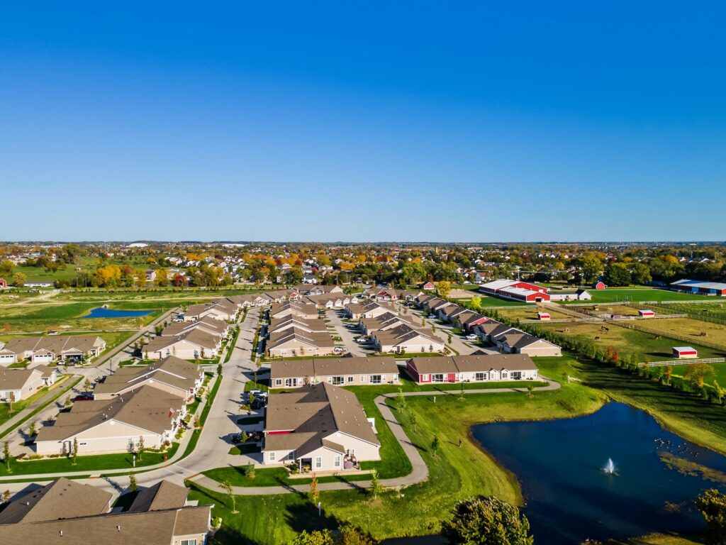 Aerial view of a suburban residential neighborhood with rows of houses, green lawns, a pond with a fountain, and farmland in the background under a clear blue sky.
