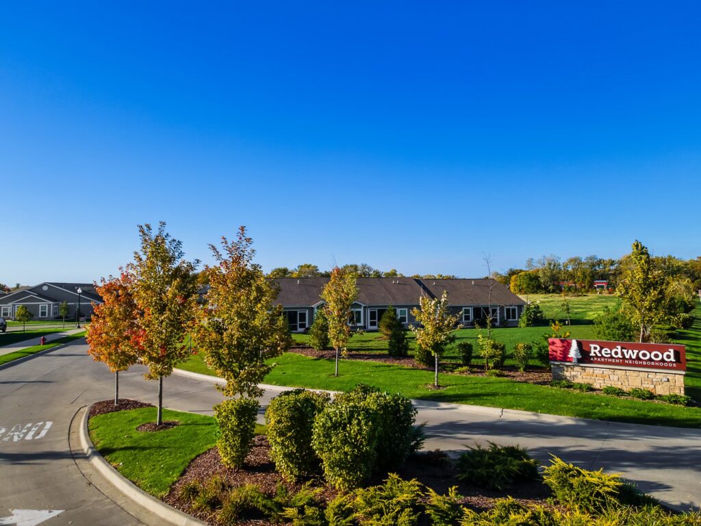 A residential neighborhood with single-story homes, landscaped lawns, young trees, and a sign reading "Redwood" under a clear blue sky.