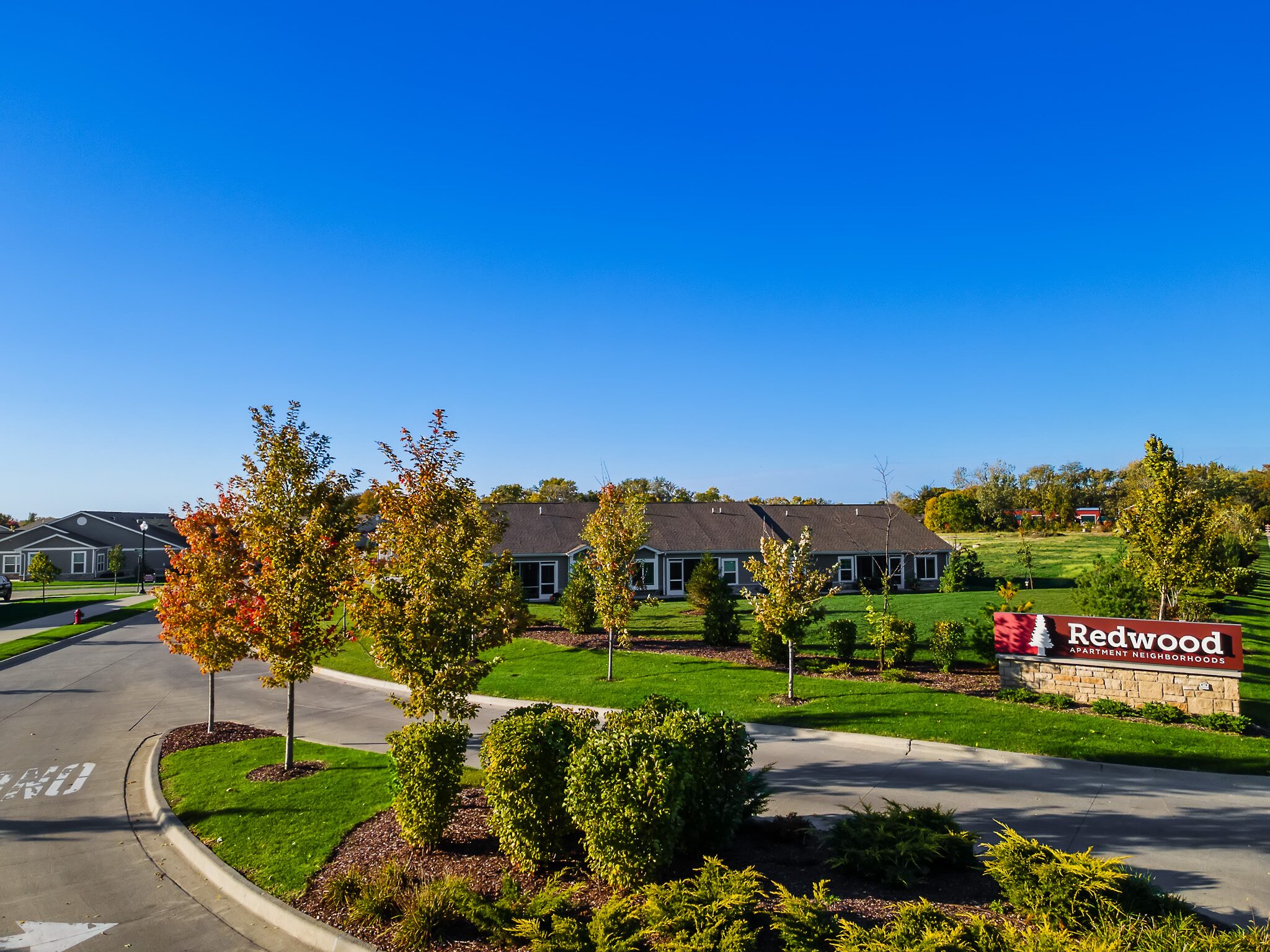 A residential neighborhood with single-story homes, landscaped lawns, young trees, and a sign reading "Redwood" under a clear blue sky.