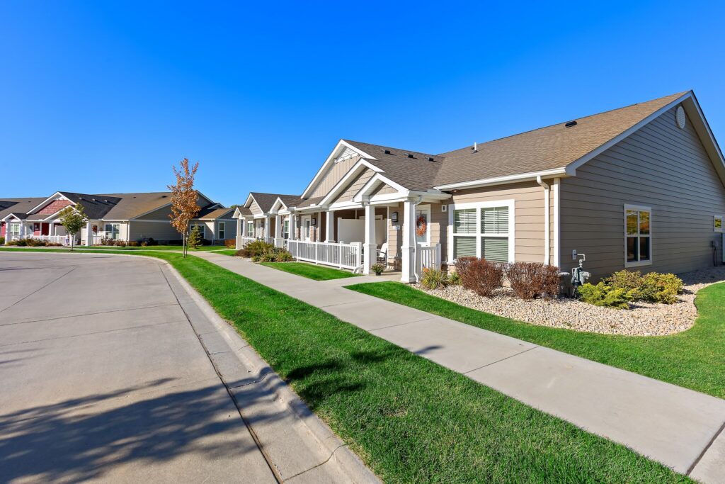 Single-story suburban houses with front porches line a clean, curved street under a clear blue sky, with well-maintained lawns and sidewalks.