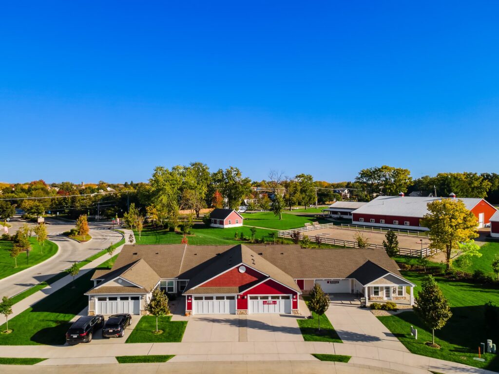 Aerial view of suburban homes with driveways and parked cars, surrounded by green lawns, trees, and red barns under a clear blue sky.