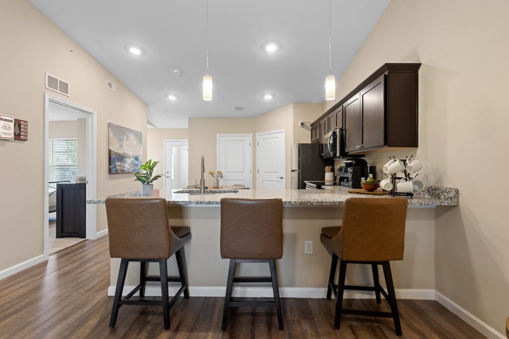 Modern kitchen with granite countertop, three brown barstools, dark cabinets, pendant lights, and wood-style flooring. Hallway and wall art visible in the background.
