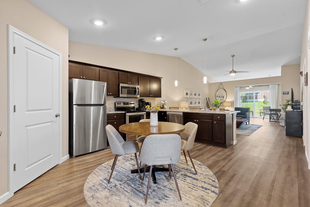 Open-concept kitchen and dining area with stainless steel appliances, dark cabinets, a round table with four chairs, and a living area in the background.