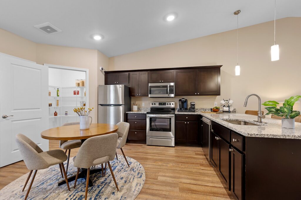 Modern kitchen with dark wood cabinets, stainless steel appliances, round dining table with four chairs, granite countertops, and a small pantry with shelves in the background.