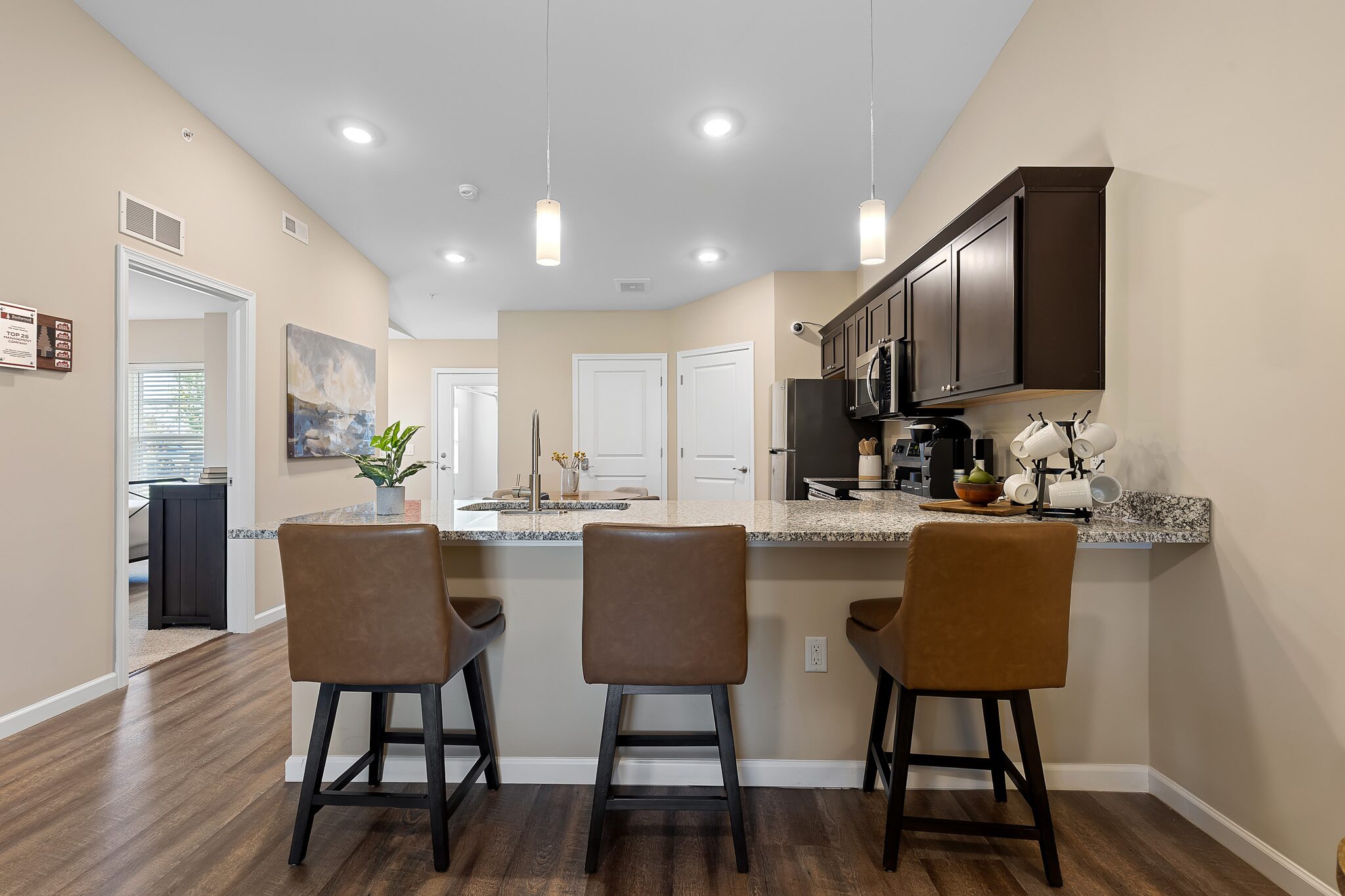 Modern kitchen with granite countertop, three brown barstools, dark cabinets, pendant lights, and wood-style flooring. Hallway and wall art visible in the background.
