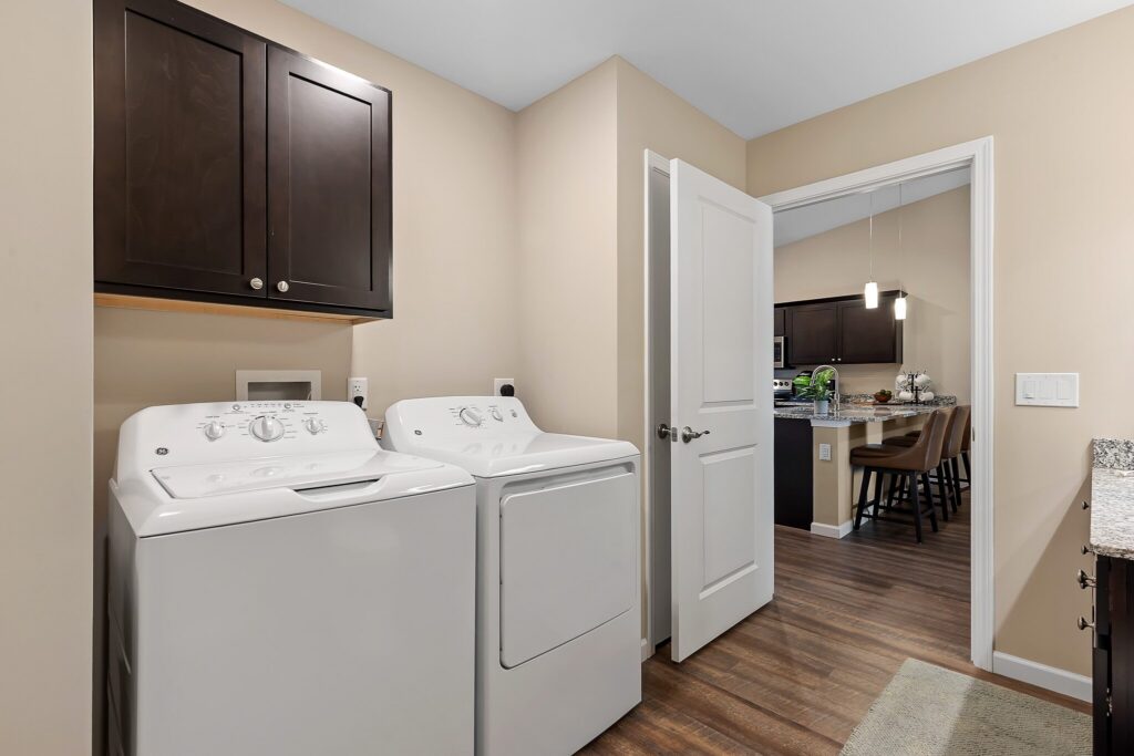 A laundry room with a white washing machine and dryer, dark cabinets above, beige walls, and an open door leading to a kitchen with wooden flooring.