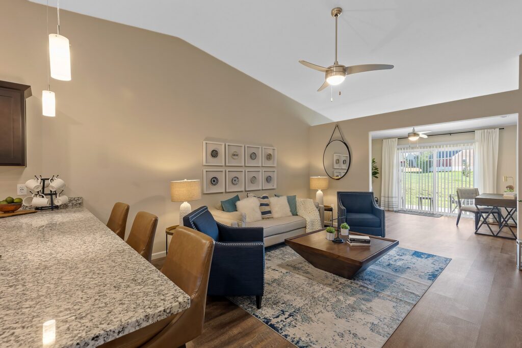 Modern open-plan living room with neutral tones, blue accents, a granite kitchen counter, and large windows leading to a patio with a view of grass outside.