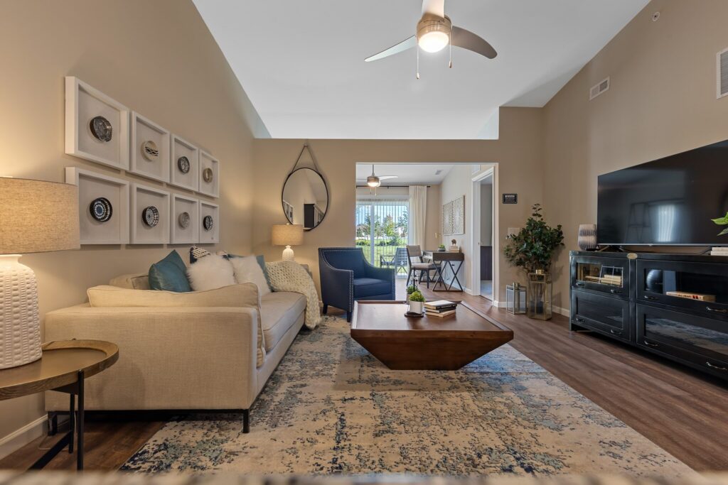 Modern living room with beige walls, a cream sofa, a wooden coffee table, wall art, a large TV, and a rug; open view to a bright room with windows and desk in the background.