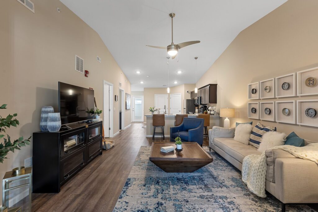 Modern living room with neutral tones, a large TV, a beige sofa, blue accent chairs, a patterned rug, and wall art, leading to an open kitchen and dining area.