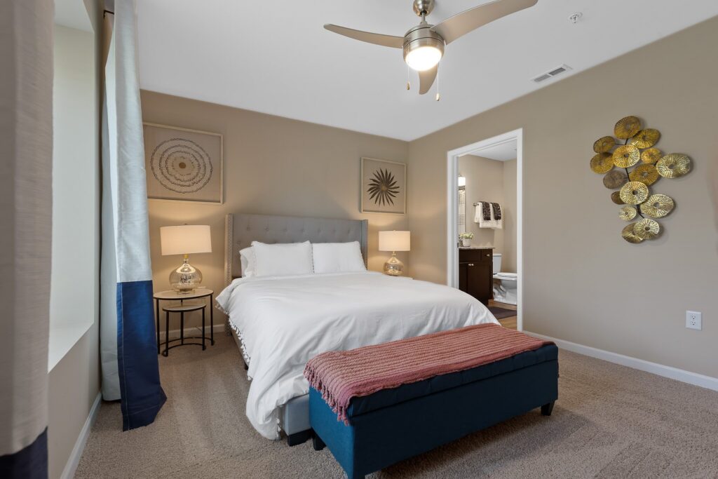 A neatly made bed with white linens and a blue bench at the foot sits in a beige bedroom with wall art, two lamps, and an open door leading to a bathroom.
