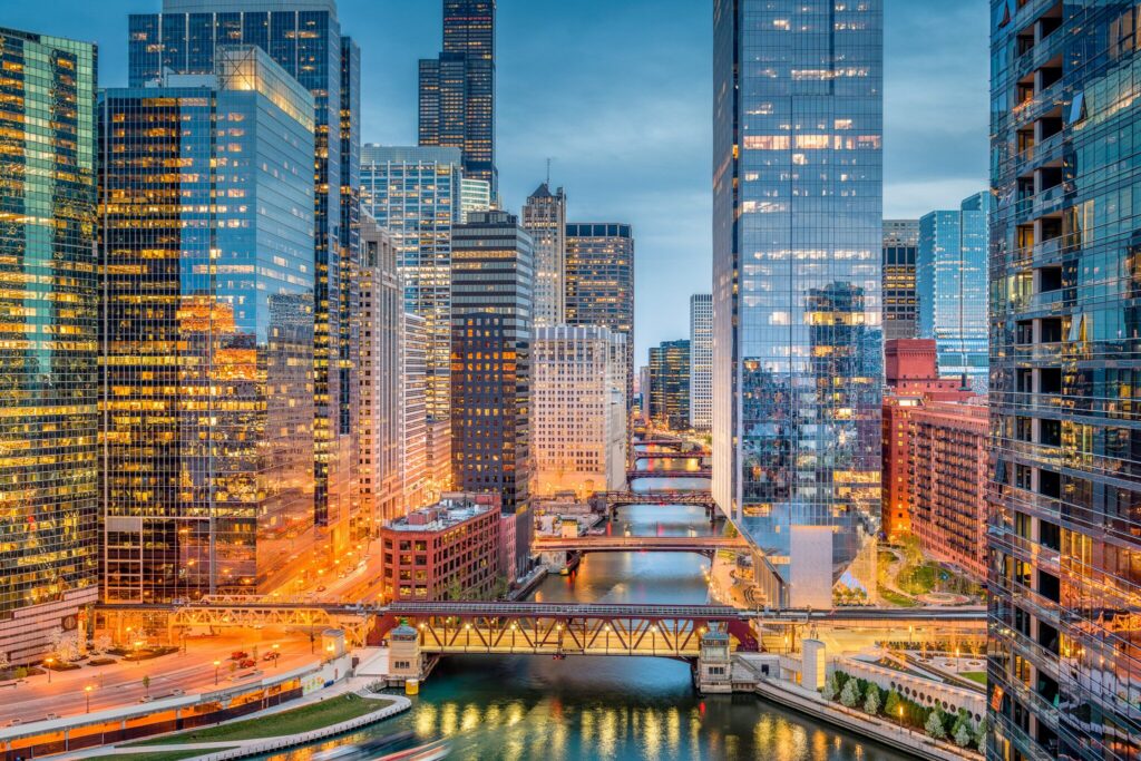 View of downtown Chicago at dusk, featuring tall glass skyscrapers, illuminated office windows, and several bridges spanning the Chicago River.