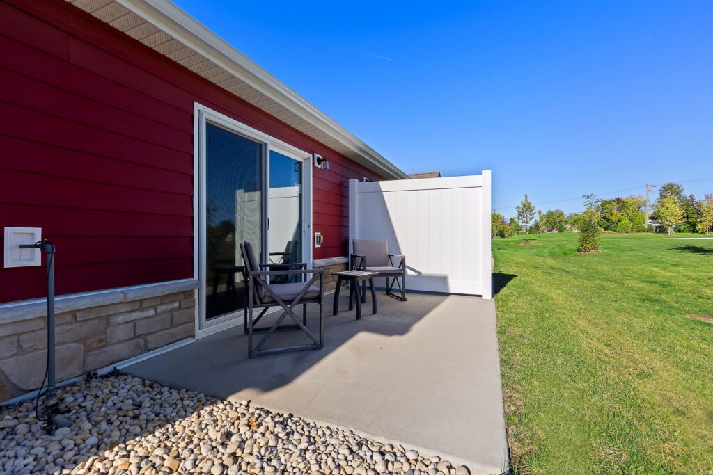 A small concrete patio with two chairs and a table outside a red house with sliding glass doors and a white privacy fence, overlooking a grassy yard.