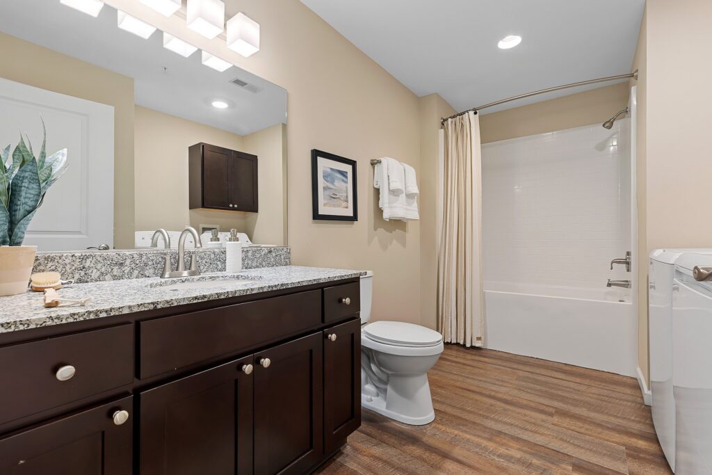 Modern bathroom with dark wood cabinets, granite countertop, toilet, bathtub with shower curtain, towel rack, and light wood flooring.