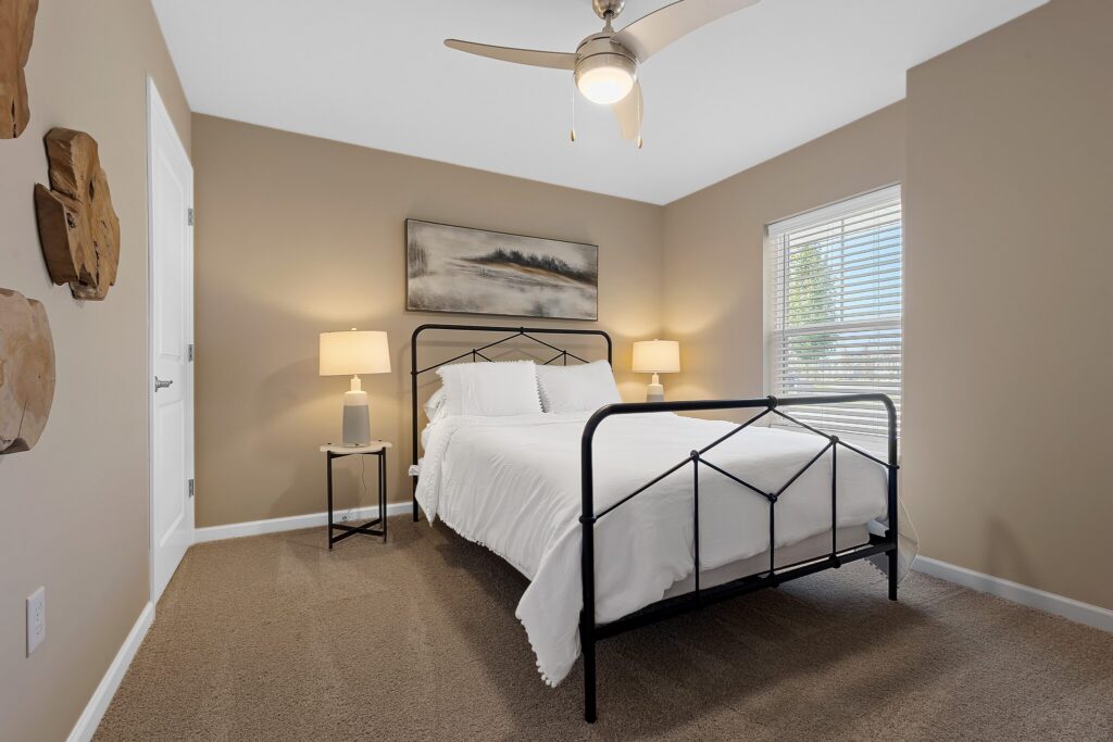 A tidy bedroom with a black metal bed frame, white bedding, two bedside tables with lamps, a wall painting, beige walls, carpeted floor, and a ceiling fan.