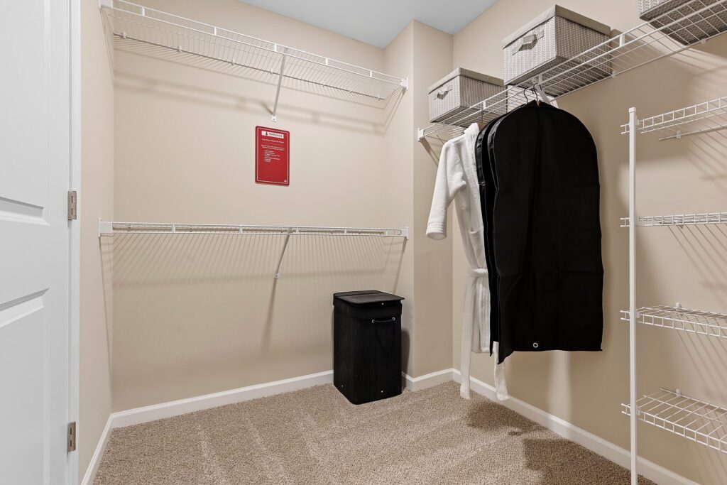 Empty walk-in closet with beige walls, wire shelving, a black hamper, storage bins, a hanging garment bag, and a white robe.