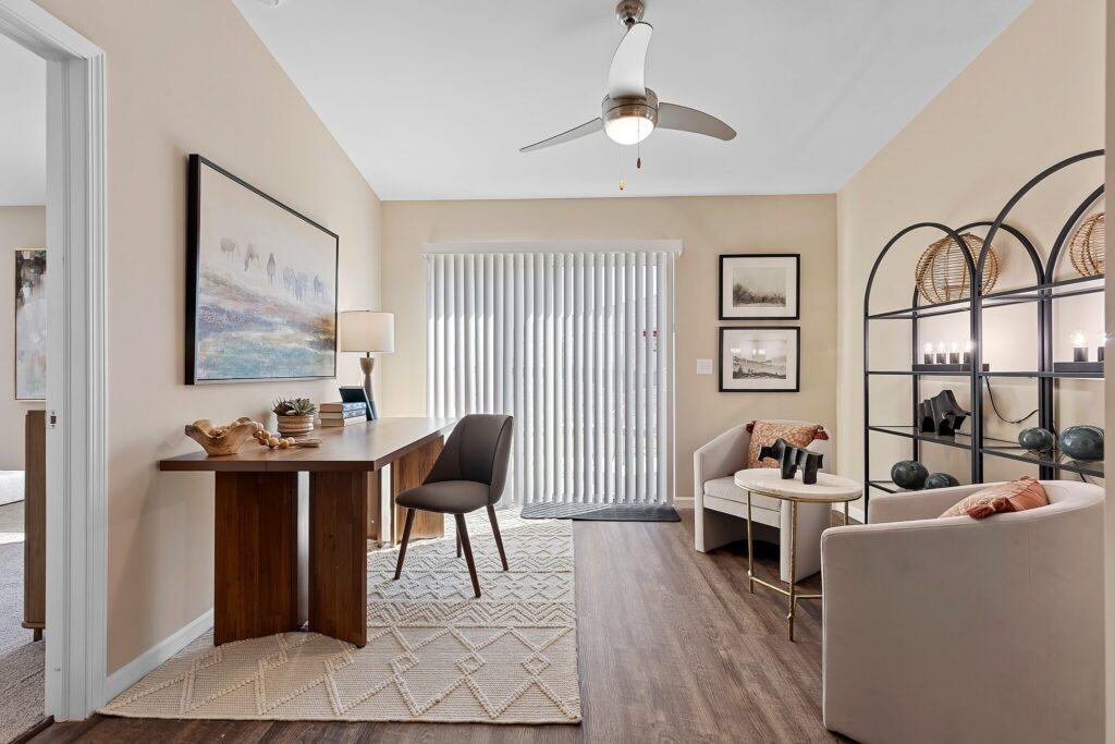 A home office with a wooden desk, chair, and lamp, a sitting area with two armchairs and a round table, shelving on the right, and blinds covering a glass door.