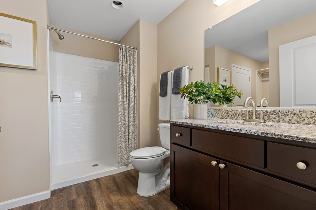 Modern bathroom with a walk-in shower, toilet, dark wood vanity with granite countertop, and potted plant on the counter. Neutral walls and wood-look flooring.