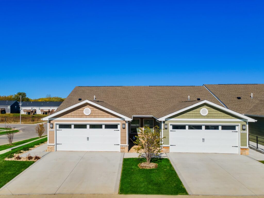 A duplex with two attached garages, beige and green siding, and a shared driveway under a clear blue sky.