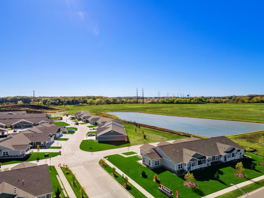 Aerial view of a suburban neighborhood with single-story houses, green lawns, and a pond under a clear blue sky.