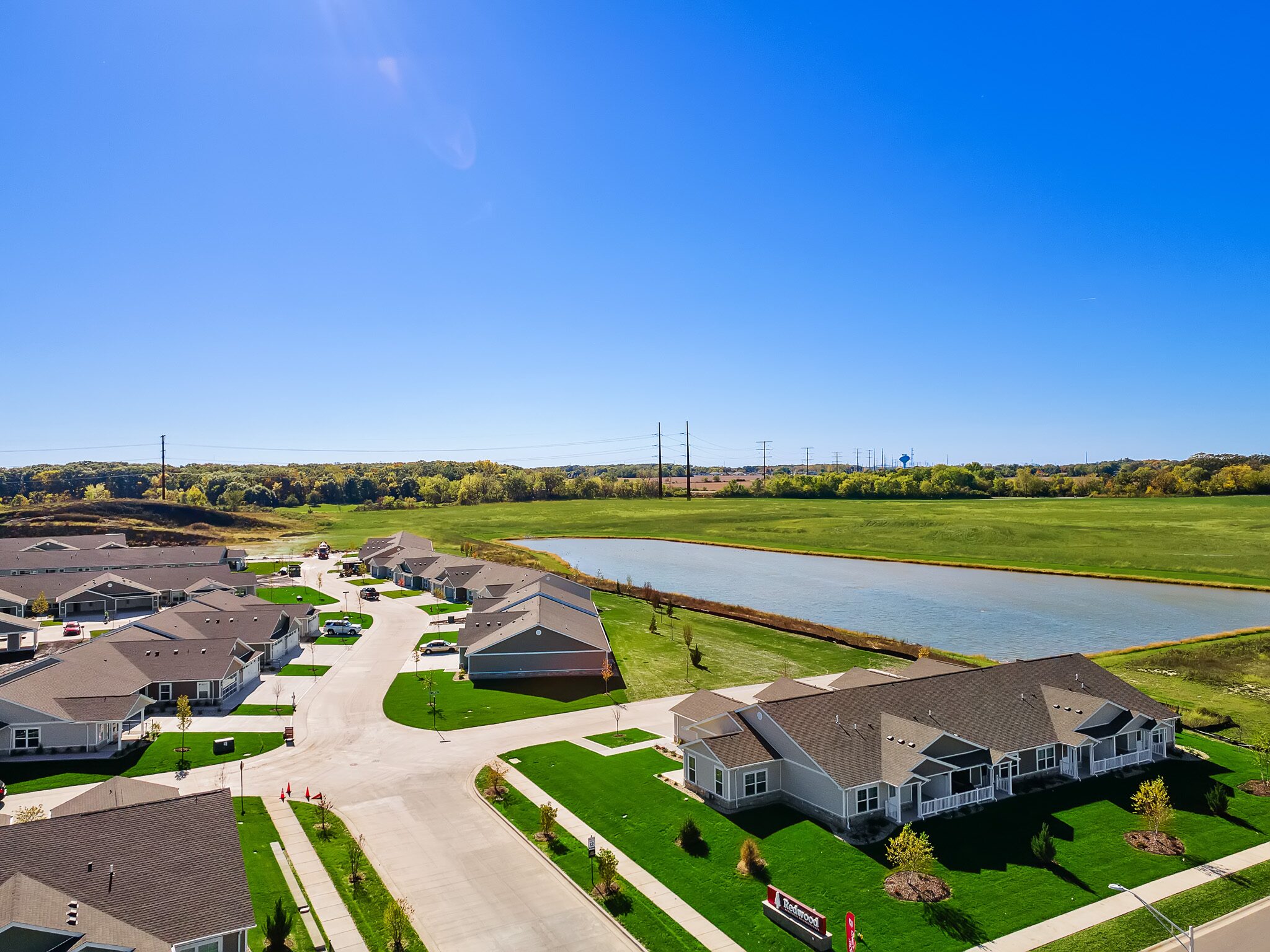 Aerial view of a suburban neighborhood with single-story houses, green lawns, and a pond under a clear blue sky.
