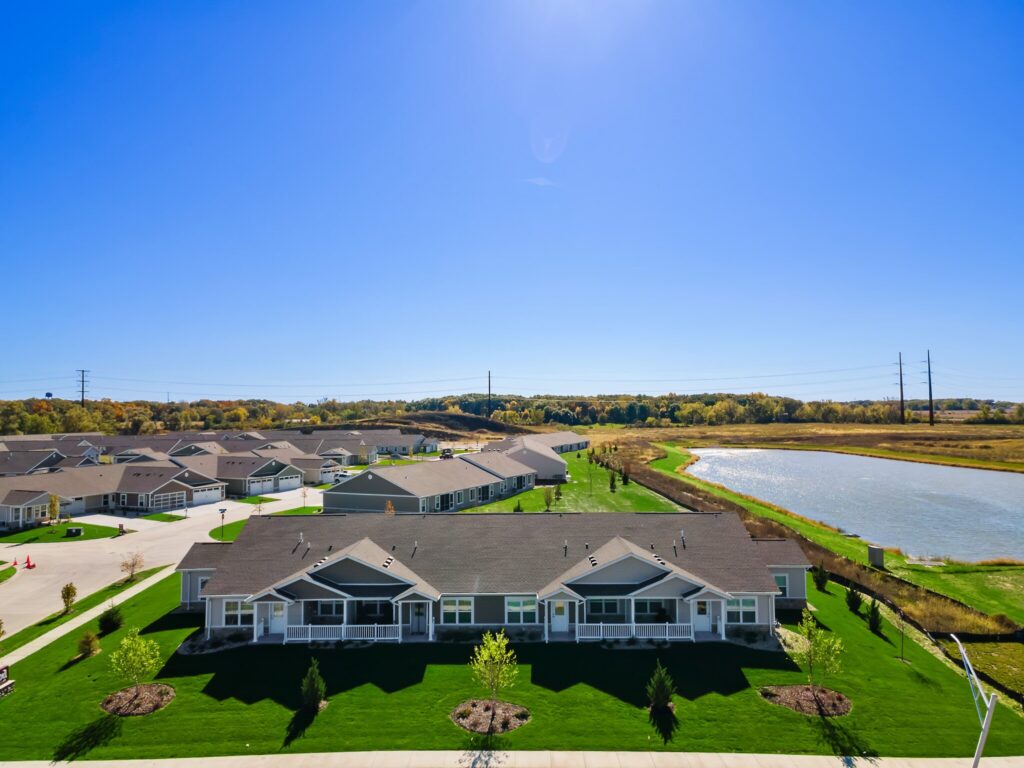 A row of single-story houses with manicured lawns faces a pond, with more houses and trees in the background under a clear blue sky.