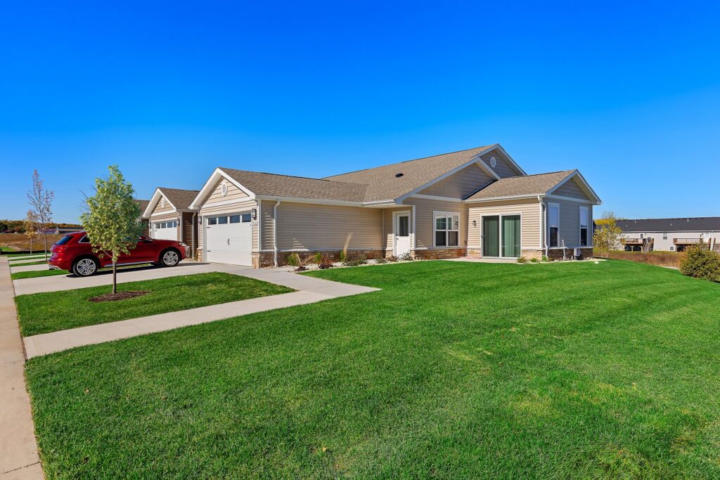 Single-story beige house with a two-car garage, red SUV parked in driveway, green lawn, and clear blue sky.