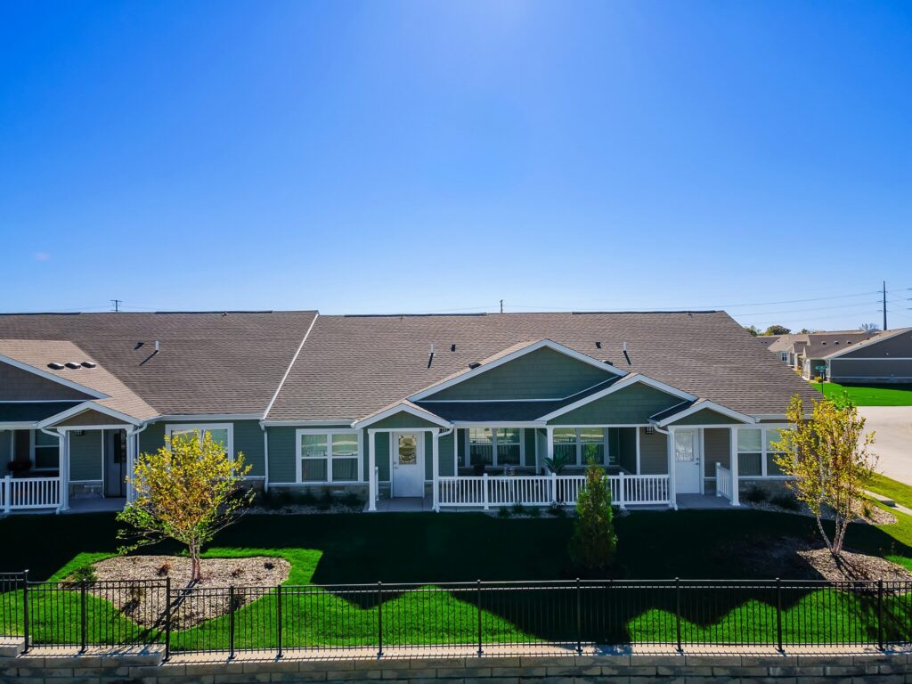 Single-story duplex with a front porch, green lawn, young trees, and a black metal fence under a clear blue sky.