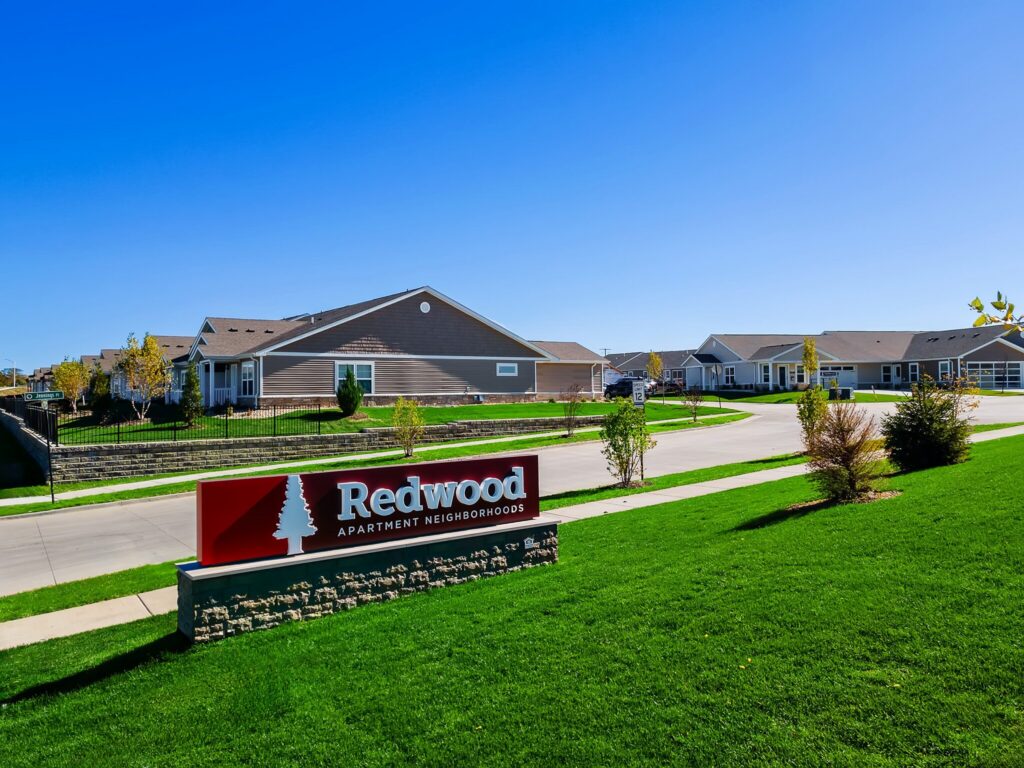 A row of single-story apartment homes with a "Redwood Apartment Neighborhoods" sign on a neatly maintained green lawn under a clear blue sky.